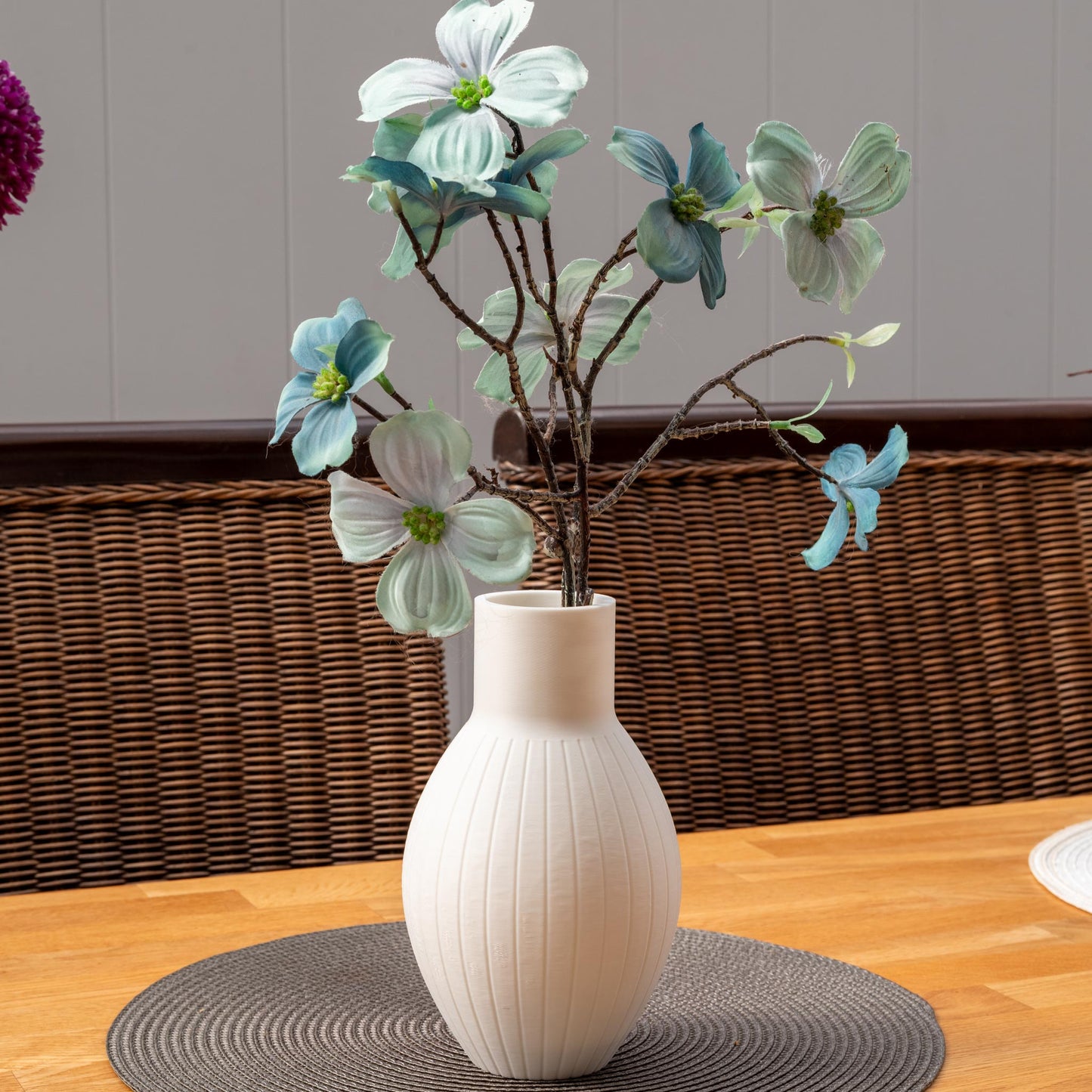 Weiße, gewellte Vase mit künstlichen Blumen in Blau und Weiß. Die Vase steht auf einem runden, grauen Tischset aus geflochtenem Material.