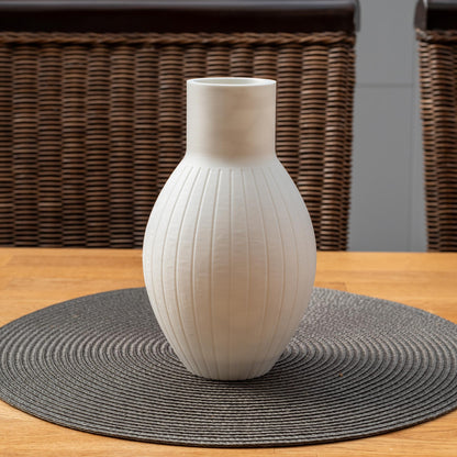 Weiße, ovaler Vase mit vertikalen Rillen, steht auf einem runden, grauen Tischset aus geflochtenem Material auf einem Holztisch.