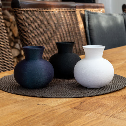 Drei nachhaltige Vasen aus PLA in Weiß, Schwarz und changierendem Dunkelblau mit matter, strukturierter Oberfläche – dekorativ arrangiert auf einer runden Platzmatte auf einem Holztisch.