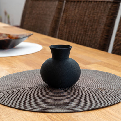 Nachhaltige, schwarze Vase aus PLA mit matter, leicht strukturierter Oberfläche in kugeliger Form – dekorativ arrangiert auf einem Holztisch mit runder Platzmatte