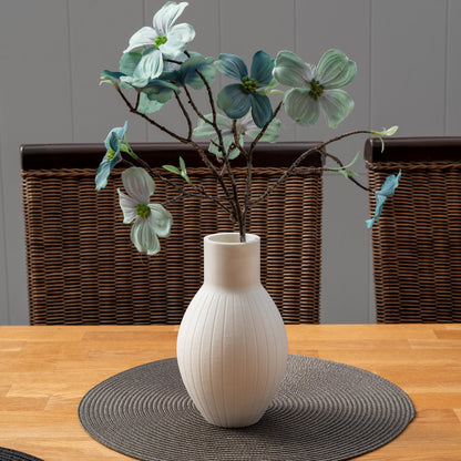 Weiße, gewellte Vase mit künstlichen Blumen in Blau und Weiß. Die Vase steht auf einem runden, grauen Tischset aus geflochtenem Material.