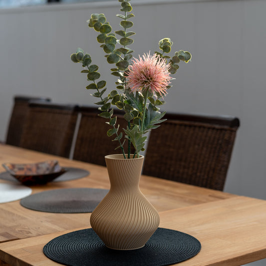 Nachhaltige Vase Line aus PLA im 3D-Druck, beige, mit gerillter Oberfläche, gefüllt mit Eukalyptuszweigen und einer rosa Blume auf einem Holztisch