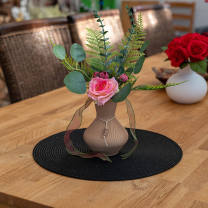Braune Vase Line aus PLA 3D-Druck mit künstlichen grünen Blättern und rosa Rose auf schwarzem Tischset auf Holztisch