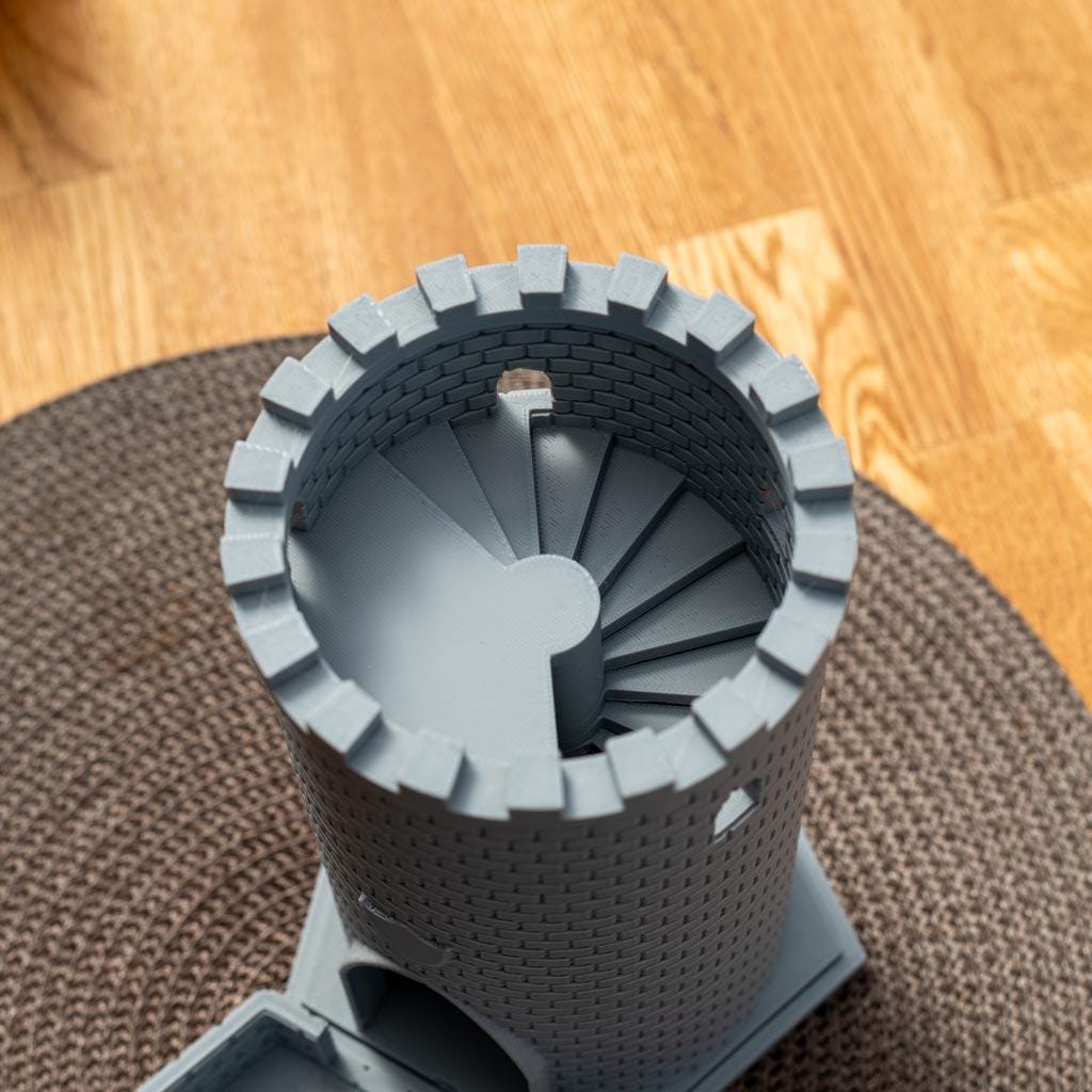 Runder grauer Würfelturm mit Zinnen und einer spiralförmigen Treppe im Inneren, auf einem Holzboden und einem braunen Teppich