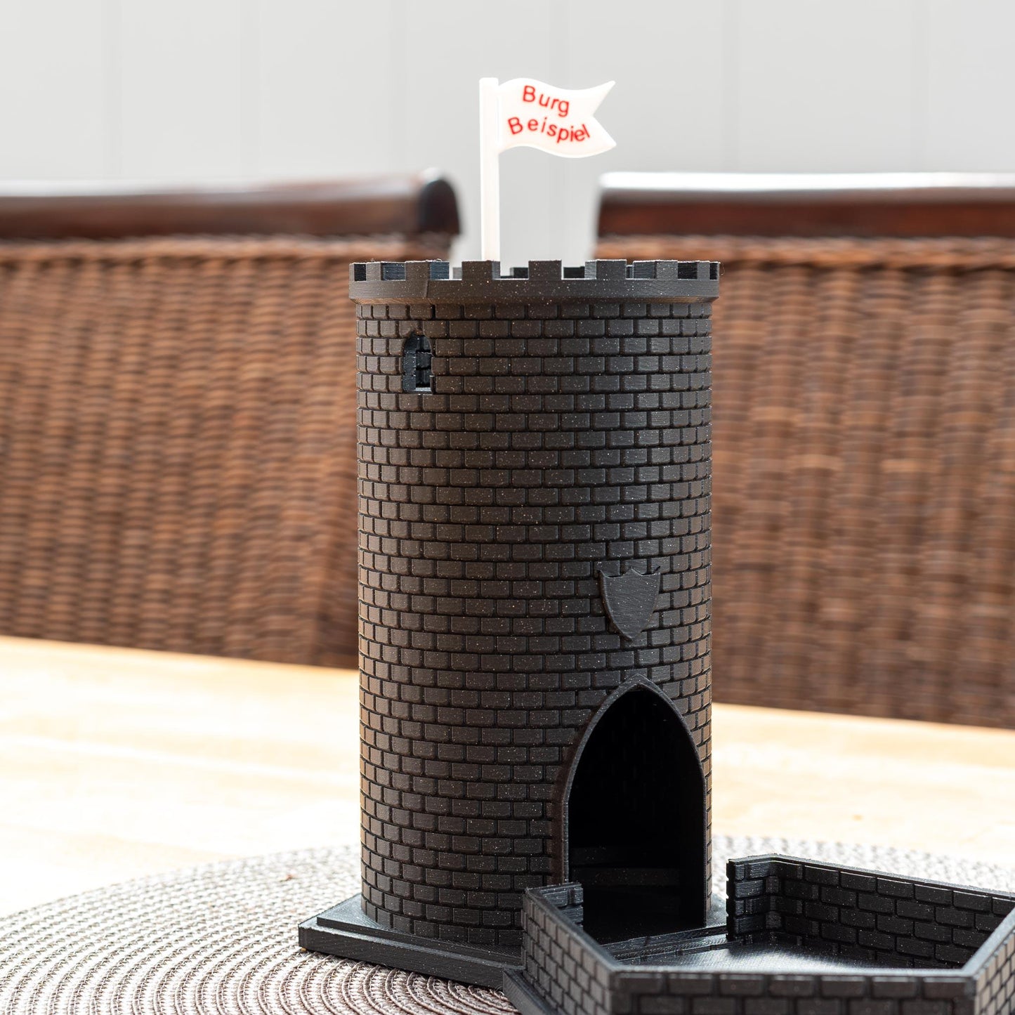 Schwarzer Würfelturm aus PLA in Form einer Burg mit Zinnen und Flagge 'Burg Beispiel' auf einem Tisch