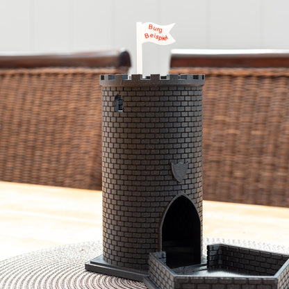 Schwarzer Würfelturm aus PLA in Form einer Burg mit Zinnen und Flagge 'Burg Beispiel' auf einem Tisch